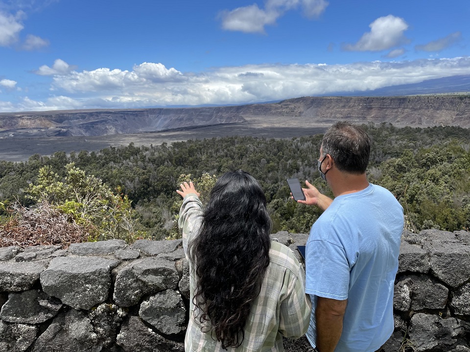 Two people standing at the rim of a vast volcanic crater under blue sky use a phone app