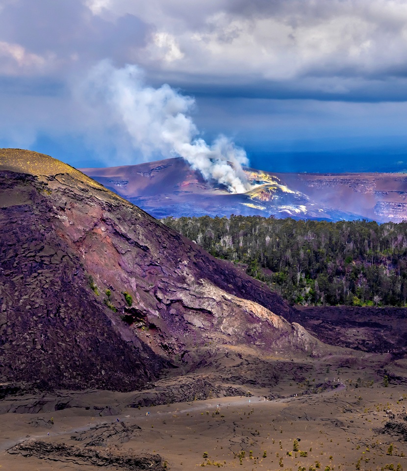 Kilauea claldera degasses in the background with Kilauea Iki crater and trail visible in the foreground with the large looming Puupuai cindercone