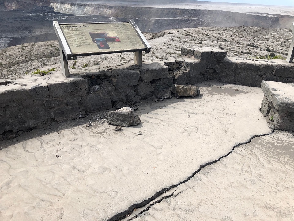 Image of concrete overlook coated in thick gray volcanic ash and a sign that reads "Fountains of Fire" on a broken rock wall. A long crack is seen on the ground