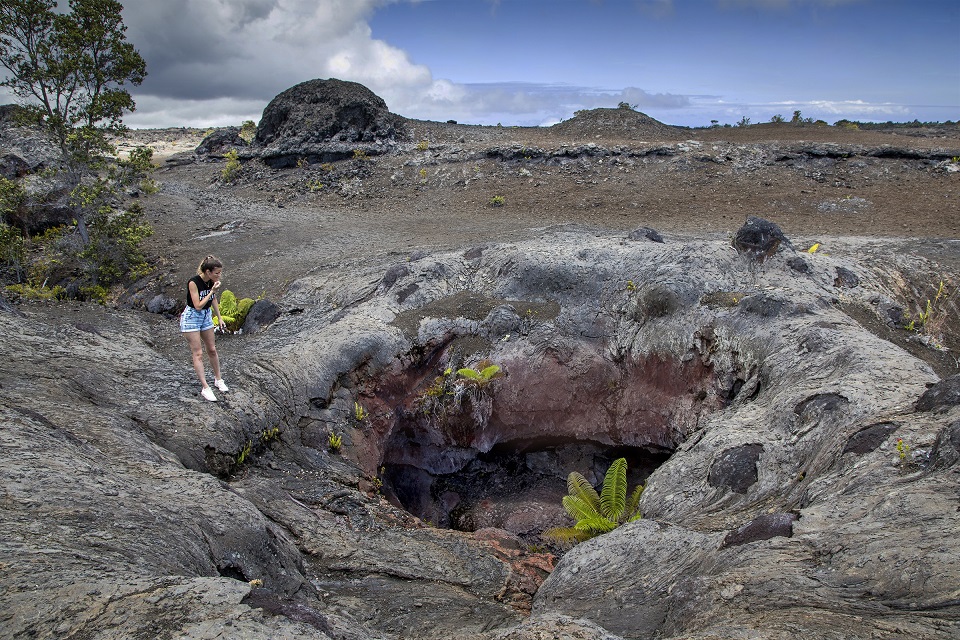 Visitor looking into fissure cave at Mauna Ulu Woman looking into volcanic cave with fern