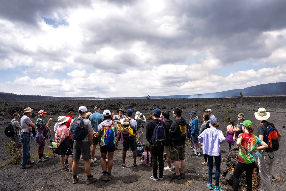 Volunteer ranger with visitors on caldera floor_J.Wei A volunteer park ranger holds a photo of an erupting volcano while standing on the caldera floor of Kilauea volcano with a few dozen visitors watching him