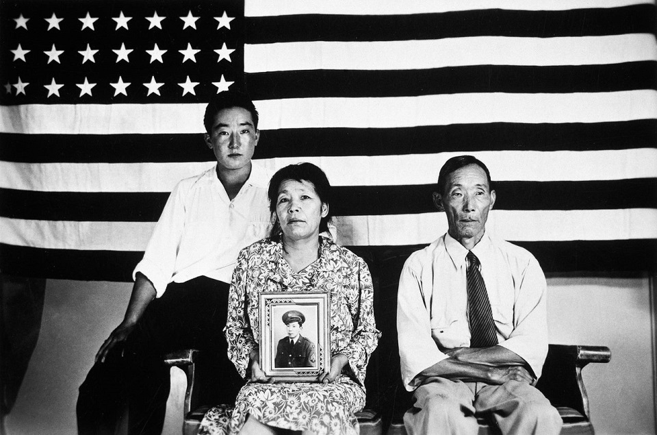 Risa and Yasubei Hirano and their son George posed in front of an American flag. Risa is holding a photograph of her son Shigera in uniform. black and white photo of Japanese American family holding photo of their serviceman son