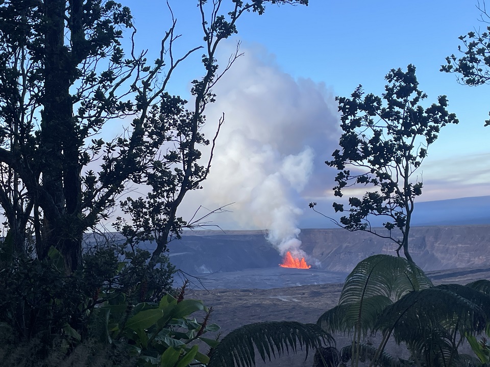 Lava fountains erupt in the distance in a volcanic crater framed by forest