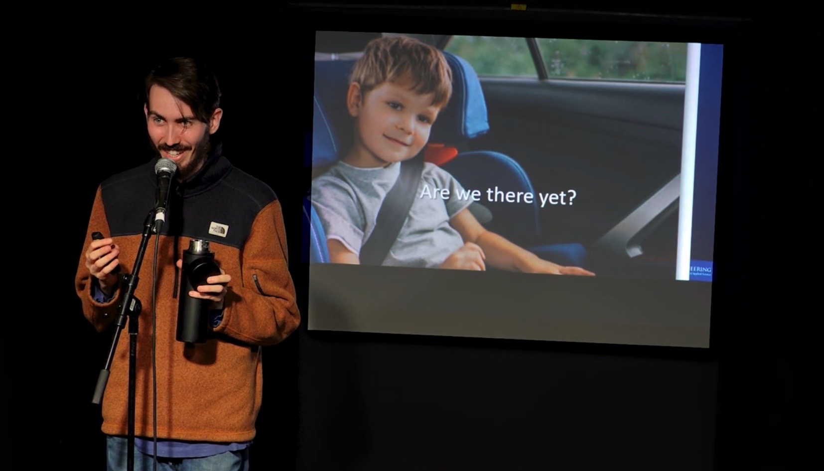 A comedian peforms on stage with a large projection screen and an image of a child wearing a seatbelt in a car. Text on the photo says "are we there yet?"