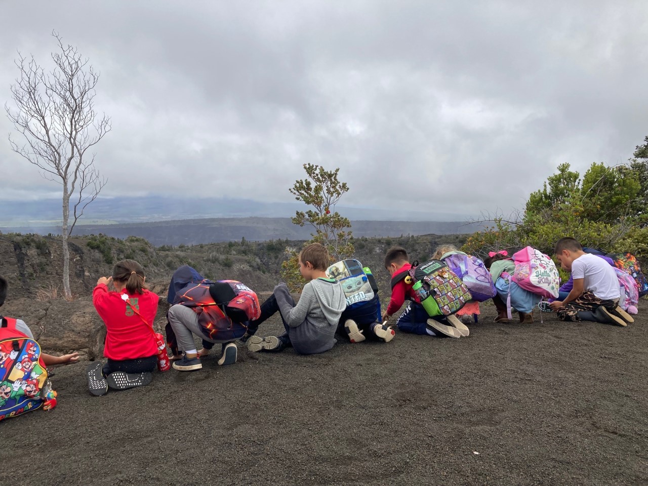 School children sit at the edge of a large gray crater