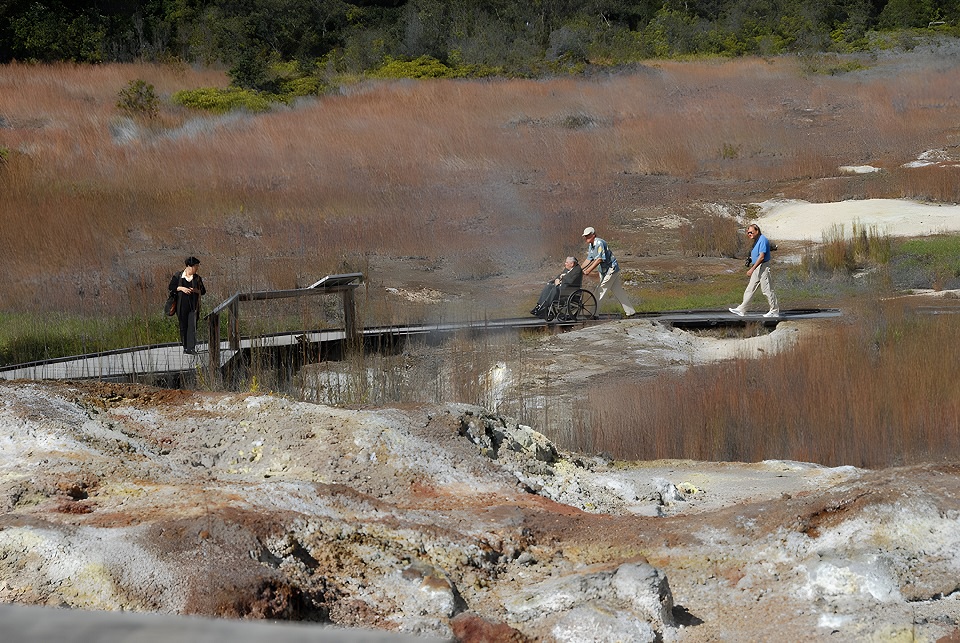 HAVO_960-Wheelchair on Sulphur Banks Trail_NPS Photo Someone pushing a woman in a wheelchair on a wooden boardwalk thorugh a steaming volcanic landscape