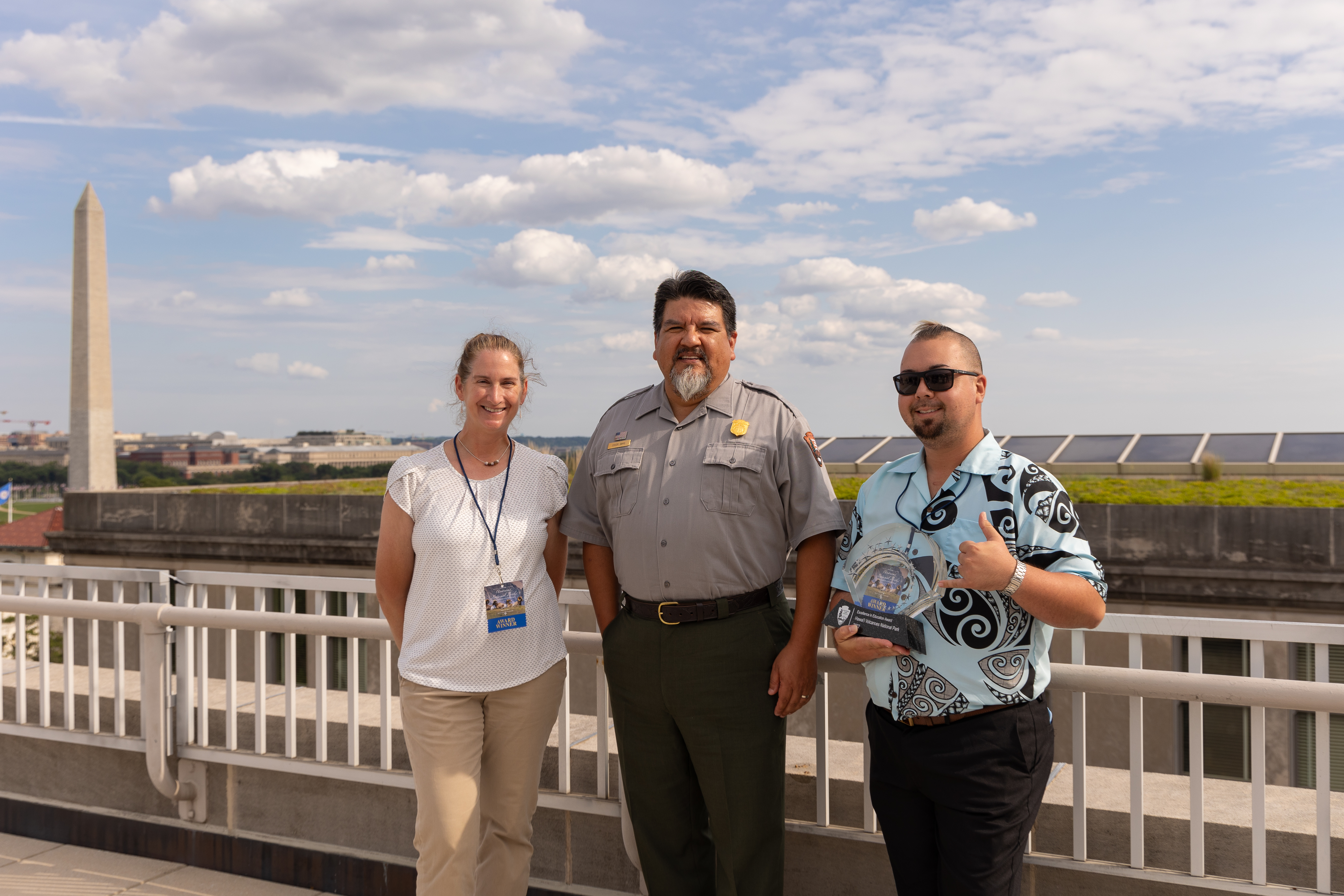 NPS Director Chuck Sams presents award Three people standing on top of a building with Washington Monument obelisk in background.