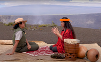 A park ranger and a hula teacher sit on a woven mat at the edge of Kilauea caldera with Mauna Loa in the background