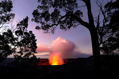 Through trees a red glow from lava illuminates the crater