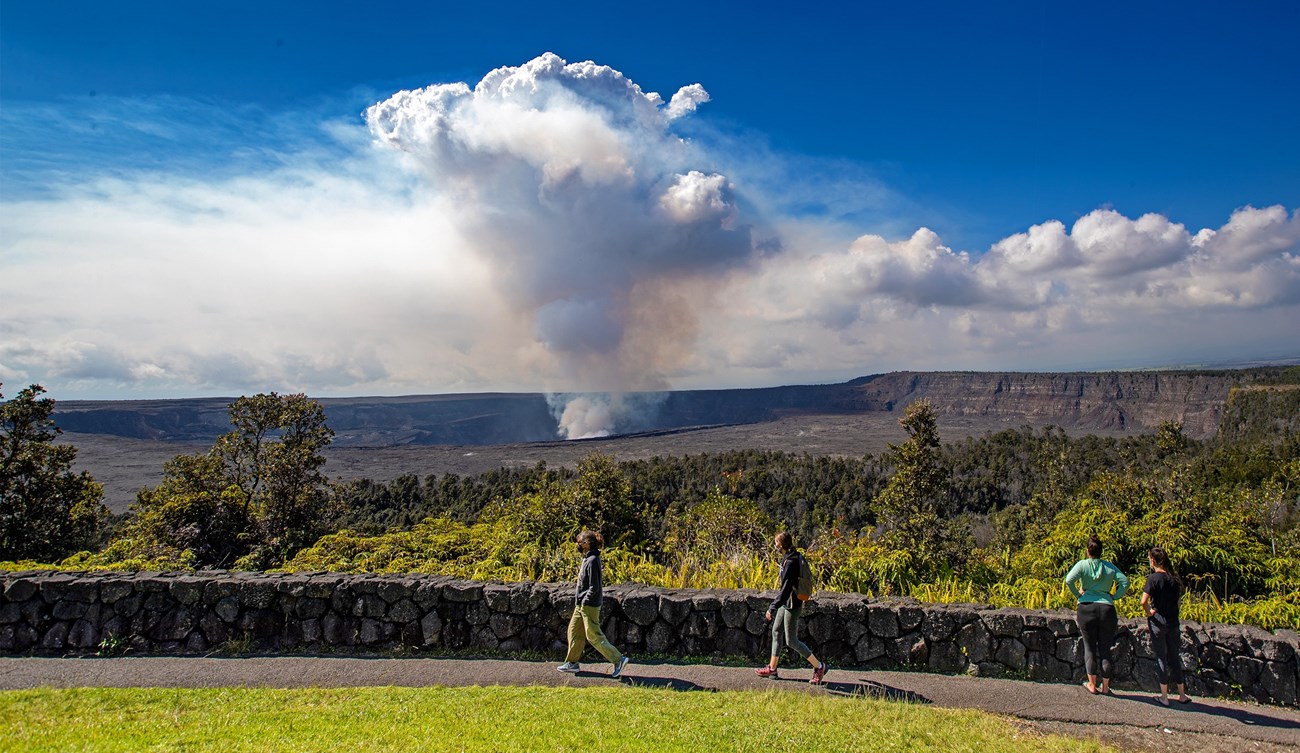 People walking on a paved path in front of a stone wall with a volcanic caldera and large steam plume beyond