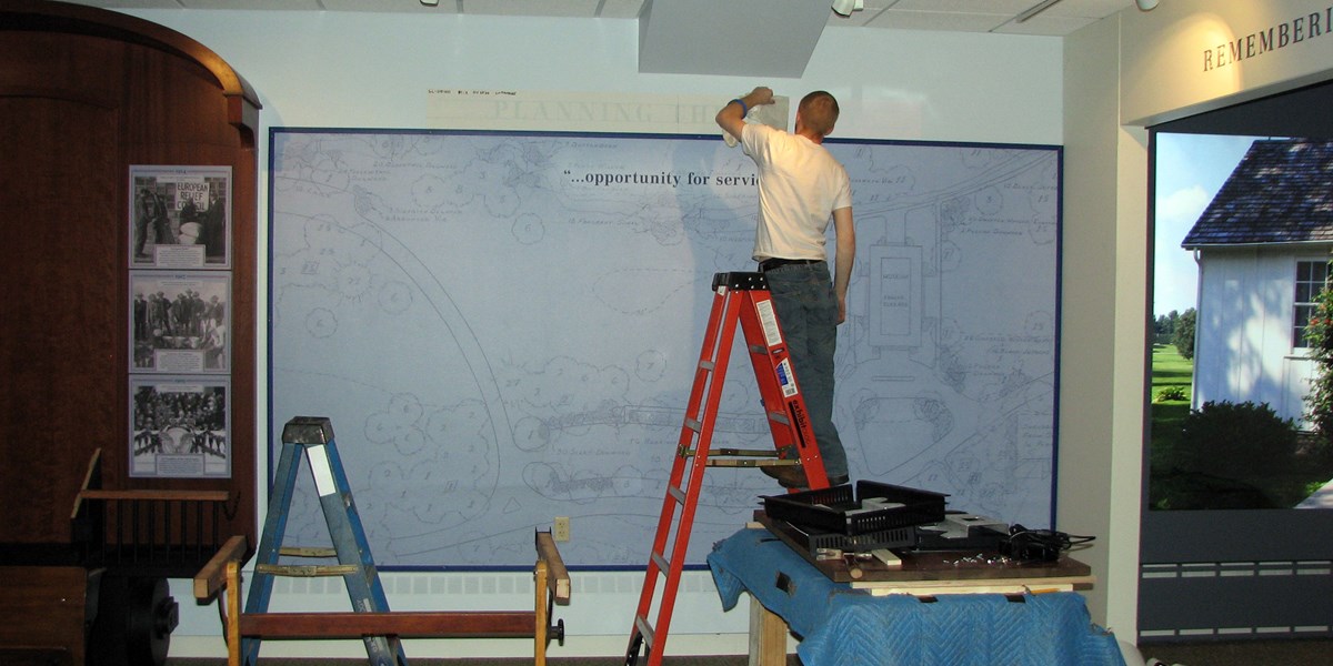 A man on a ladder stencils a heading on a museum exhibit under construction. A man on a ladder stencils a heading on a museum exhibit under construction.