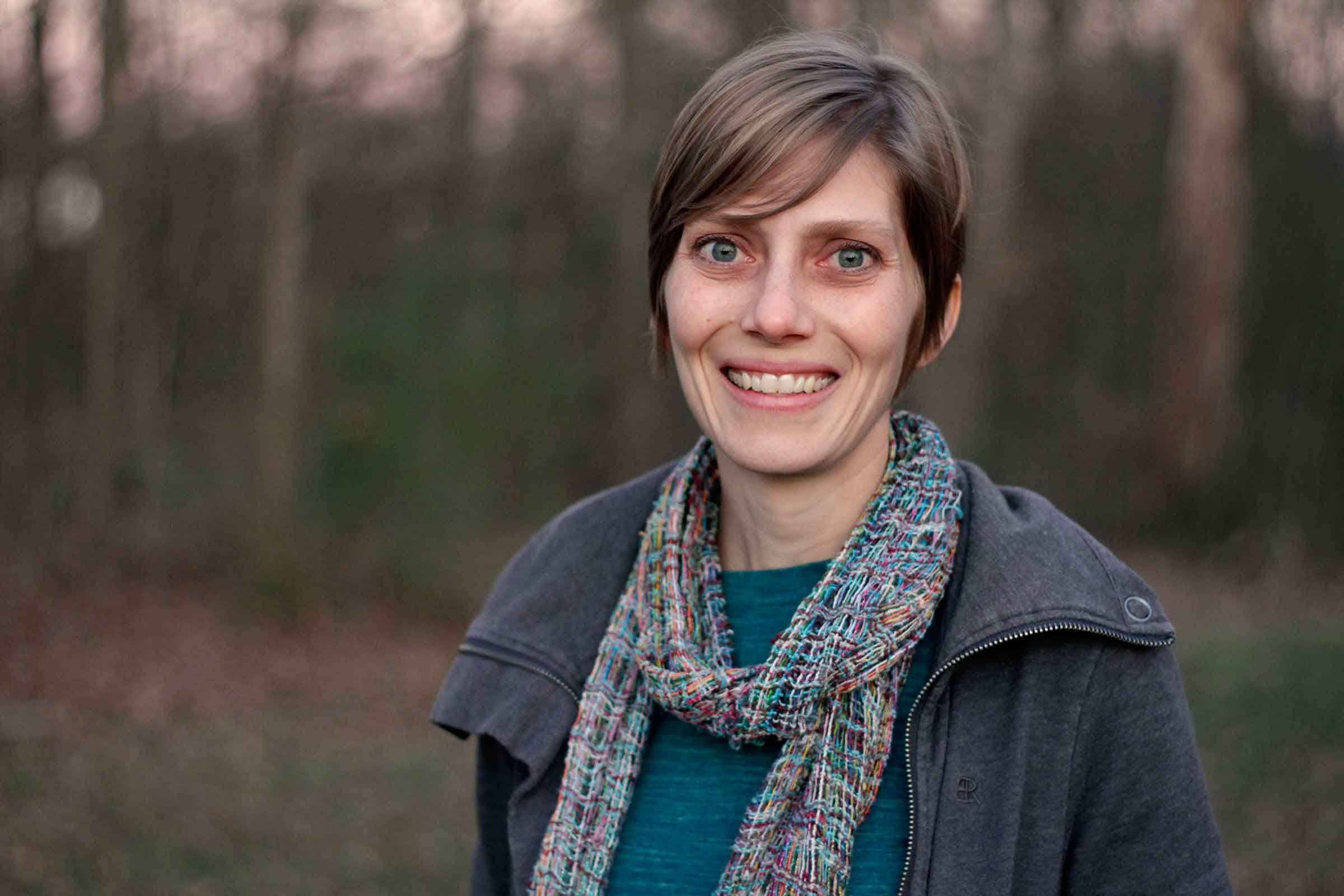 A woman in a sweater and scarf smiles in a wooded setting.