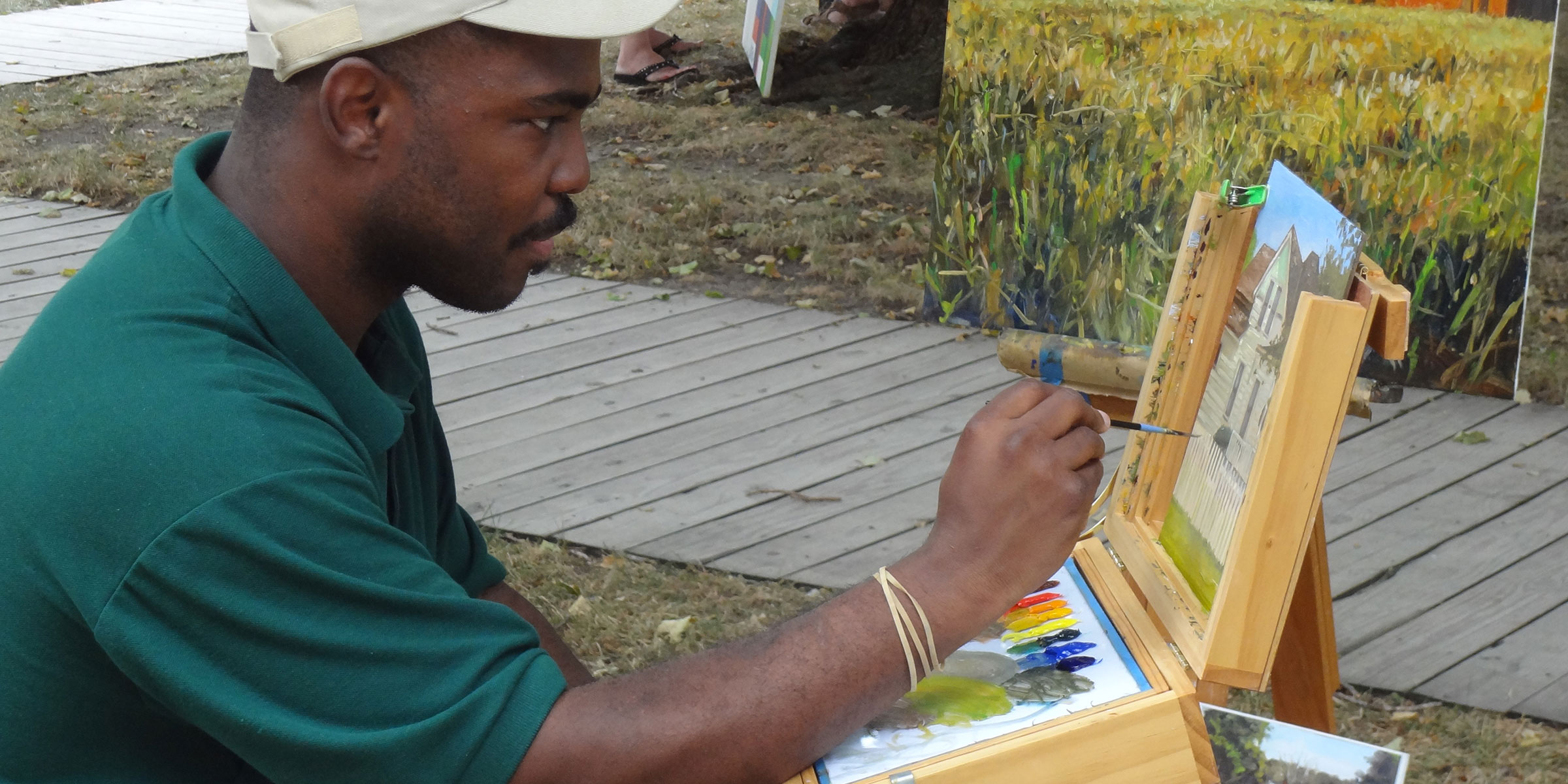A man seated at an easel paints and displays finished paintings. A man seated at an easel paints and displays finished paintings.