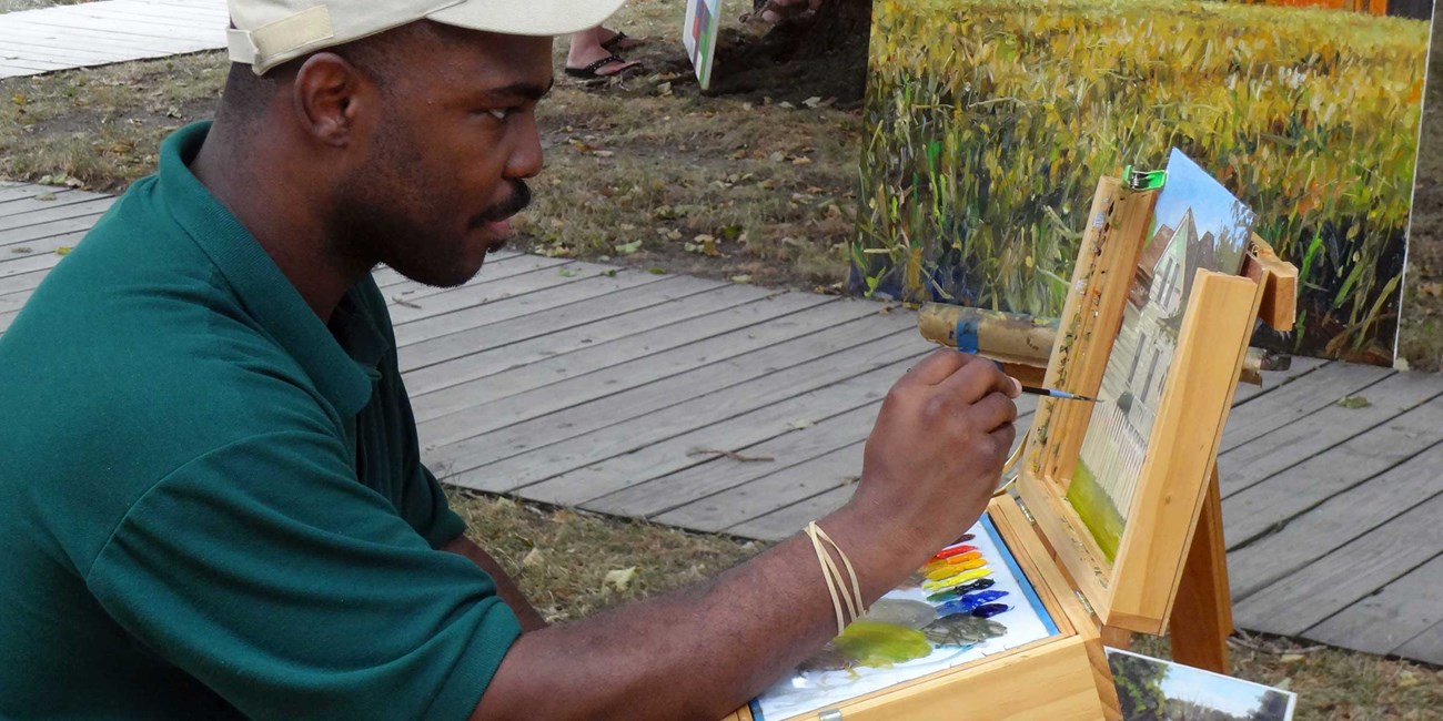 A man seated at an easel paints and displays finished paintings.