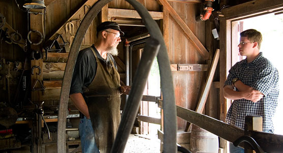 A blacksmith chats with a visitor inside a blacksmith shop.