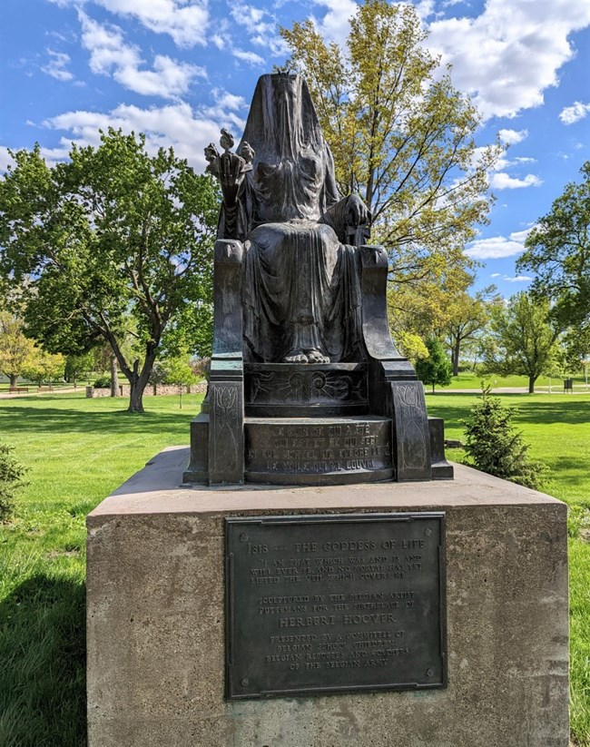 A bronze statue of a seated goddess is displayed in a park.