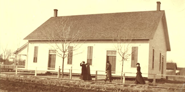 Men and women in conservative 1800s dress walk to a simple meetinghouse.