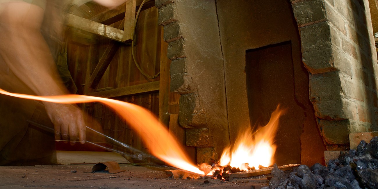 A man with tongs holds metal in a forge with bright orange flames.