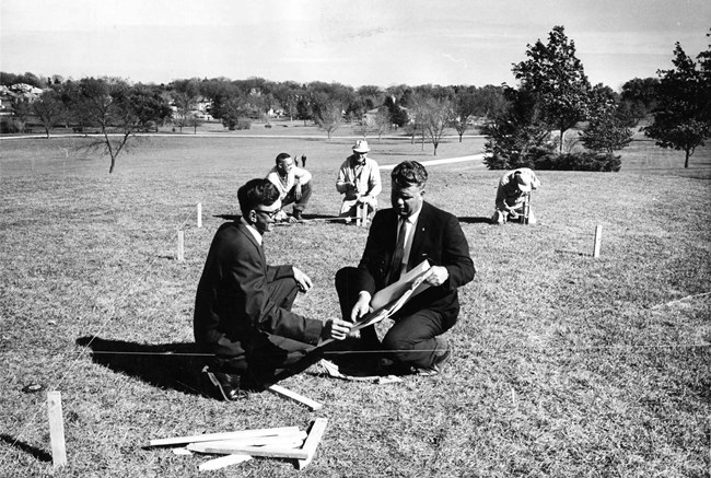 Two men kneel in the grass and study blueprints as three other men stake out a plot with string.
