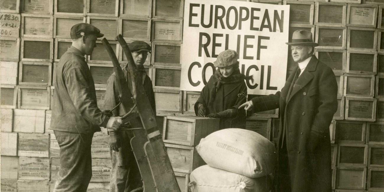 A man in a suit and hat stands with three relief workers with sacks of flour.