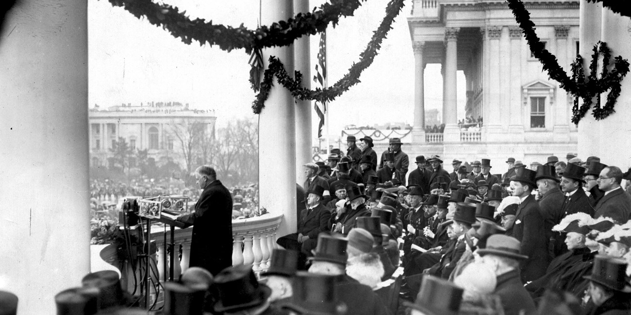 Dignitaries in formal suits and top hats surround Herbert Hoover as he speaks among the pillars of the White House.