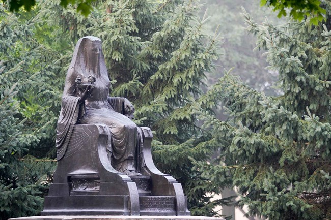 A bronze statue of a seated goddess is displayed in a park.