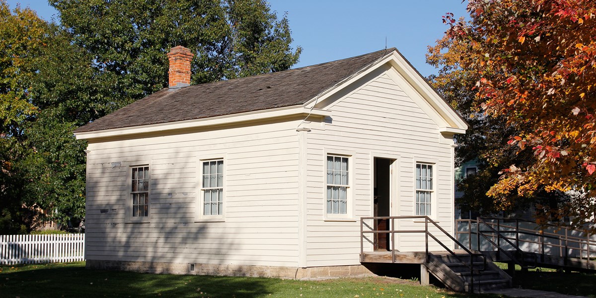 A one-story wooden building painted off-white has two windows and a central doorway. A one-story wooden building painted off-white has two windows and a central doorway.