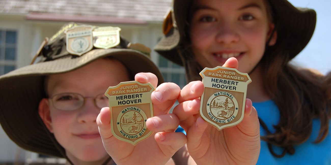 Two smiling kids hold junior ranger badges up to the camera in front of a cottage.