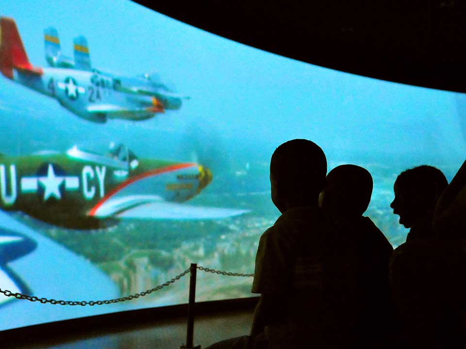 Rise Above Panoramic Movie Silhouetted children watch World War Two era airplanes on a wraparound screen in a darkened theater.