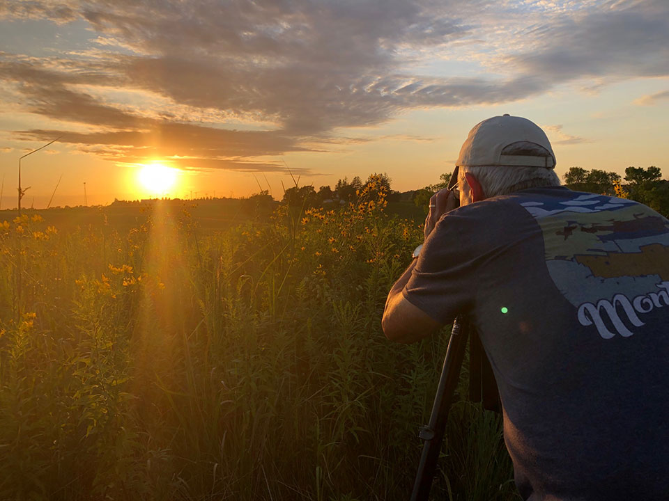 2019 Artist-in-Residence Jim Schlett A photographer leans into his camera tripod to photograph a prairie sunset.