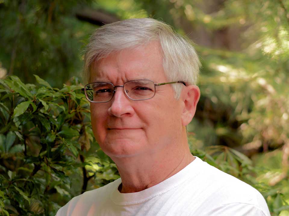 Photographer Jim Schlett A man with gray hair and glasses poses with his arms crossed in an outdoor natural setting.