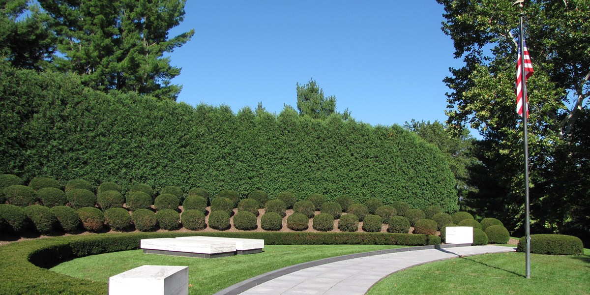 Two plain marble ledgers mark graves in a semicircular planting of landscape shrubs and a United States flag. Two plain marble ledgers mark graves in a semicircular planting of landscape shrubs and a United States flag.