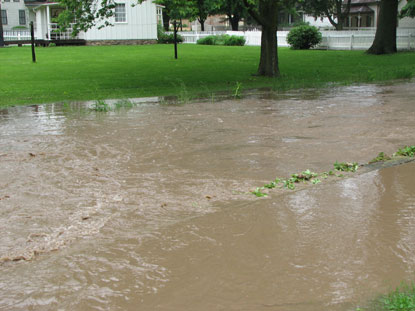 High, muddy water tops the banks of a creek near a building.