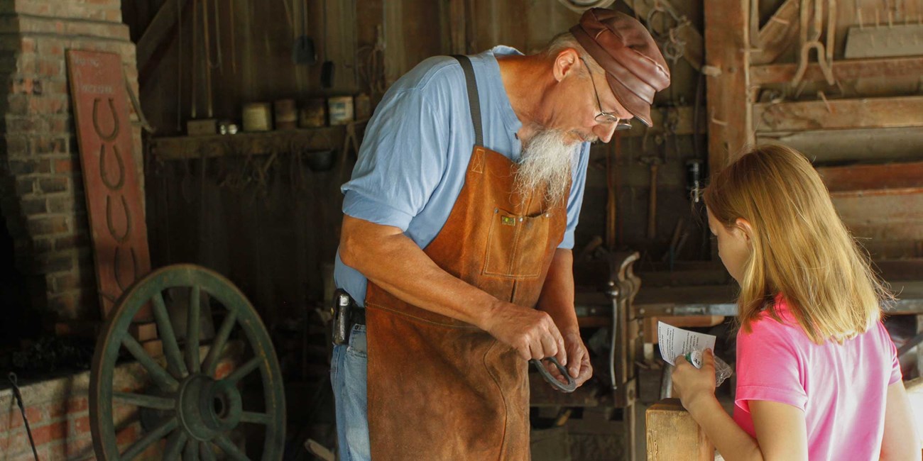 A blacksmith in an 1870s style stop shows a horseshoe to a little girl.