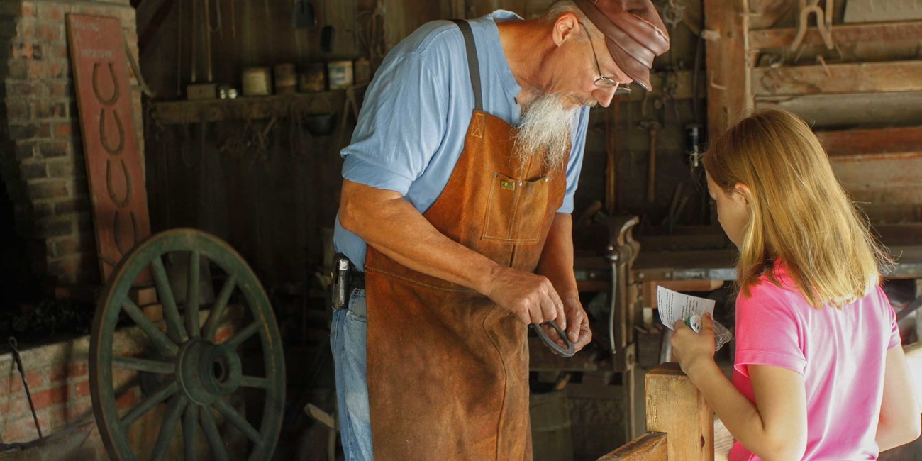 A blacksmith in a traditional shop shoes a horseshoe to a girl.