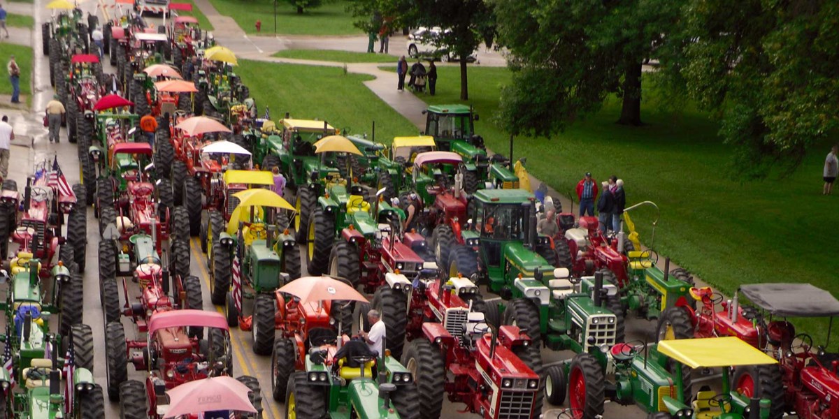Rows of colorful antique tractors line up for a parade on a street near a park. Rows of colorful antique tractors line up for a parade on a street near a park.