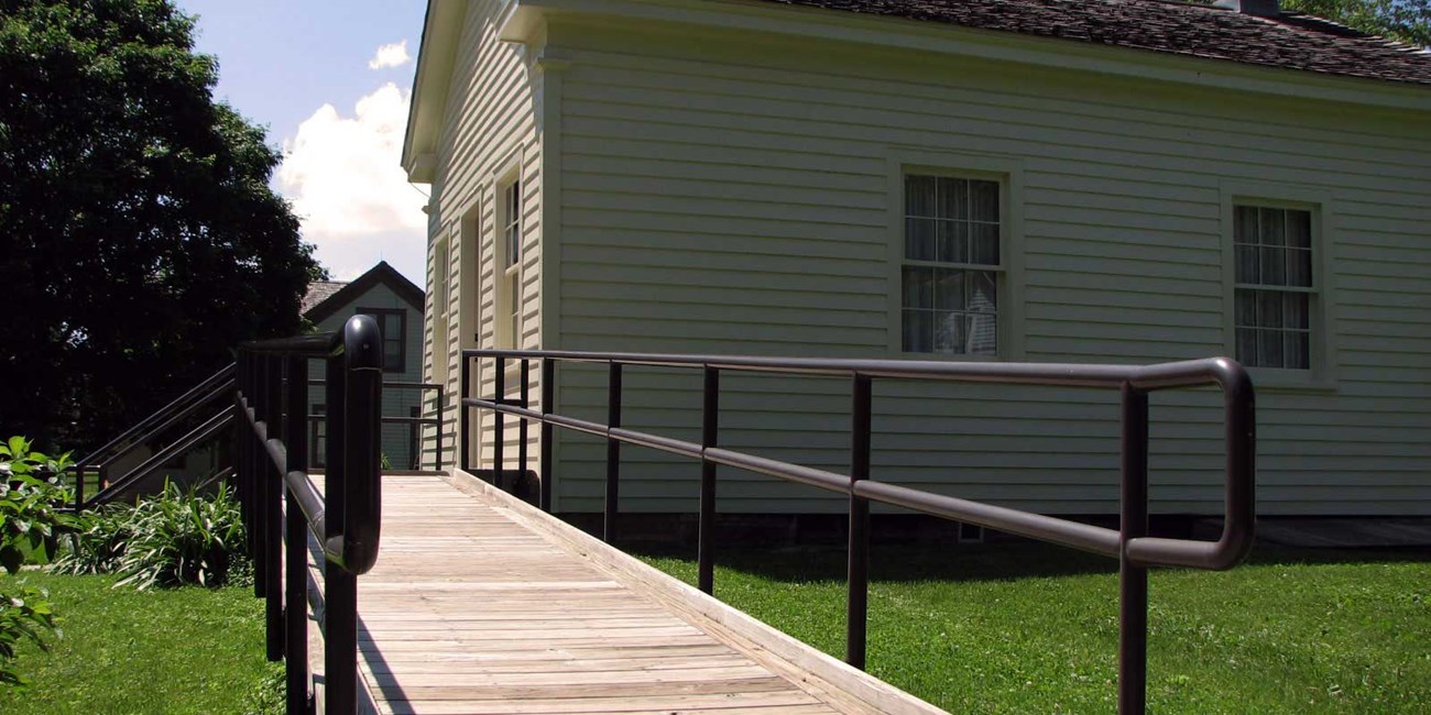 A wooden ramp with a metal railing leads to the front door of an old schoolhouse.