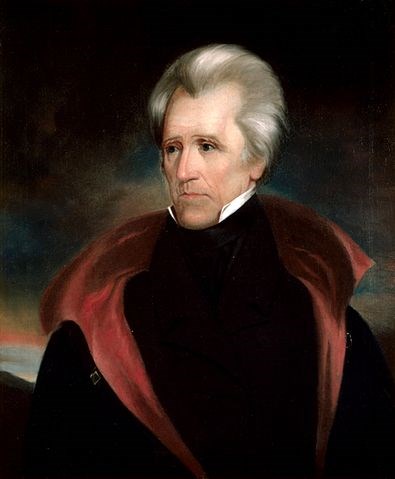 portrait of man with grey hair from stomach up wearing a cloak type jacket with red collar/lining and blacvk shirt covering neck, white collar showing just above the neck line