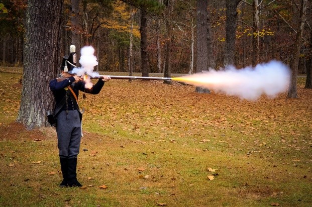 a man in period military clothing shooting a musket, smoke and flame coming from pan and barrel