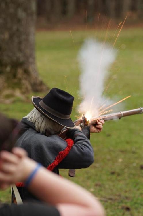 Image displays a living history event volunteer wearing darkly colored clothing, firing a musket rifle. Sparks and smoke are coming from the gun.