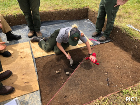A person on their hands and knees using a trowel to dig in a certain spot while others stand nearby