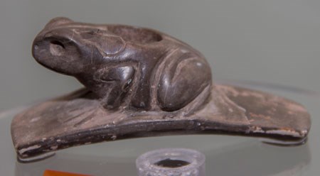 Frog Pipe A dark-colored stone pipe with a curved, flat base and a cutout of a frog atop it with the head to the left and rear to the right