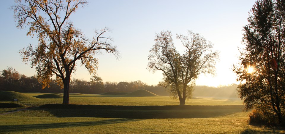 The rising sun peeks through the trees over top of grass-covered mounds