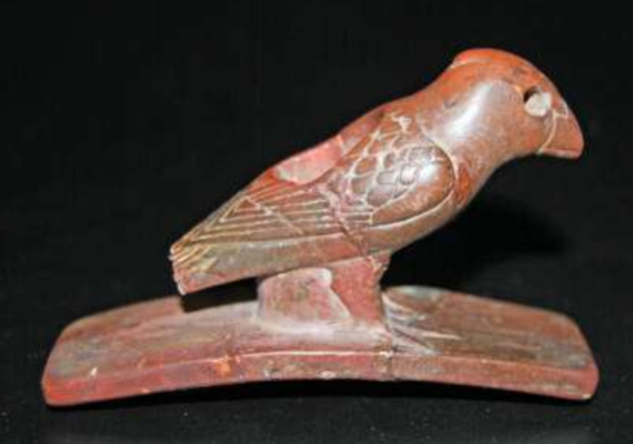 Pipestone Bird A red-colored stone pipe with a curved, flat base and a cutout of a bird atop it with the head to the top right and tail to the bottom left