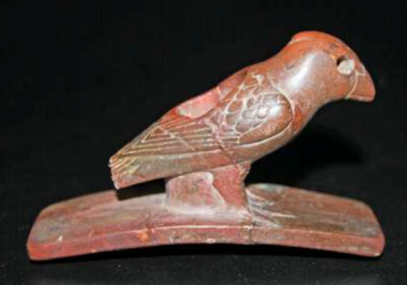 A red-colored stone pipe with a curved, flat base and a cutout of a bird atop it with the head to the top right and tail to the bottom left
