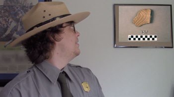 A ranger in a flat hat looks up to his left at a piece of broken pottery in a photo with 837 written below it