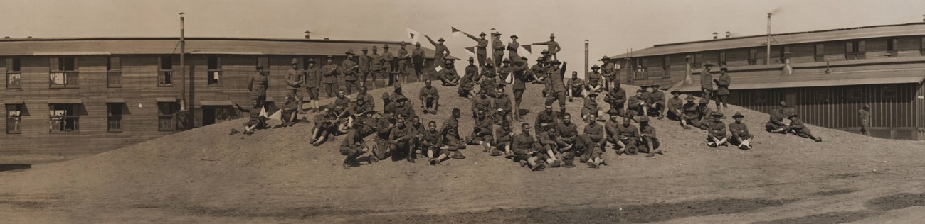 Several men in uniforms standing and sitting atop a mound of earth between two large buildings