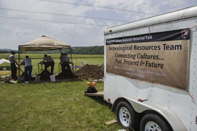 Several people working under a tent with a travel trailer in the foreground