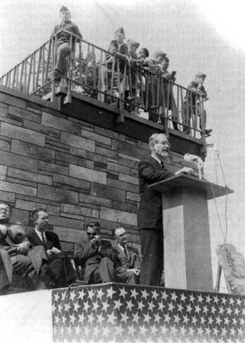 A man speaks at a podium with children on the building roof looking down