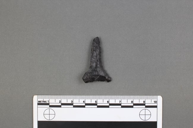 lt= black tiangular shaped stone drill in center of photo with ruler bordering the bottom for size comparison.